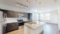 This single-floor residence showcases a contemporary open-concept design that seamlessly connects the kitchen and living areas. The kitchen is a standout feature, equipped with dark espresso cabinetry, a white subway tile backsplash, and a full suite of stainless steel appliances including a range, microwave, refrigerator, and dishwasher. A central island with a granite countertop and undermount double sink serves as both a functional workspace and a natural gathering point, illuminated overhead by elegant cylindrical pendant lights. A track lighting system provides additional task lighting along the kitchen ceiling.

The living area flows naturally from the kitchen, bathed in natural light from multiple windows fitted with horizontal blinds that offer views of the surrounding exterior. Light wood-style flooring runs throughout the main living spaces, adding warmth and continuity to the design.

The bedrooms feature plush carpeting and ceiling fans, with windows that allow ample natural light. One bedroom includes direct access to the main living area. The bathroom is clean and functional, featuring a soaking tub with a tiled surround, a curved shower rod, and wood-style flooring. Storage closets are also visible within the unit, contributing to its practical layout.