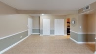 Located at 1 Lakeside Rd in Greenville, South Carolina, this single-floor home offers a well-organized and cohesive layout with consistent design elements throughout. The interior is defined by neutral two-tone wall treatments — warm tan on the upper half and a muted sage-gray on the lower — complemented by crisp white trim and baseboards. Light beige tile flooring runs throughout the main living areas, providing a clean and durable foundation.

The galley-style kitchen is a standout feature, showcasing rich, warm-toned wood cabinetry with raised-panel detailing, granite-patterned countertops, a stainless steel sink, and black appliances including a refrigerator and range. The layout maximizes counter and storage space on both sides of the corridor, making it highly functional.

The open living and flex areas are generously proportioned, offering flexibility for a variety of uses. Natural light filters in through multiple windows fitted with horizontal blinds, providing pleasant outdoor views of the surrounding greenery. Several built-in closets with bifold doors provide ample storage throughout the space.

The bathroom includes a dark-toned vanity with a white countertop and sink, a large mirror, and overhead lighting. Additional storage is accessible via a walk-in closet area adjacent to the bathroom. The overall ambiance is clean, neutral, and move-in ready.
