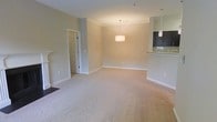 This single-floor residence showcases an open-concept layout that seamlessly connects the main living area to the kitchen. The living space is finished with plush neutral-toned carpeting and warm beige walls, creating a calm and inviting atmosphere throughout. Crown molding lines the ceiling, adding a touch of architectural elegance to the room. A centrally hung pendant light fixture serves as a focal point, providing warm ambient lighting to the open space. The kitchen is partially separated by a raised bar-height counter finished with granite countertops, offering a practical surface for casual dining or meal preparation. Dark cabinetry contrasts attractively against the lighter countertop and wall tones, lending the kitchen a modern, polished look. Recessed lighting in the kitchen area further enhances its functionality. A doorway visible from the main living area leads to additional spaces within the unit, suggesting a well-considered floor plan. White trim and baseboards throughout provide clean, crisp definition between the walls and flooring. Overall, the space presents a comfortable, move-in-ready environment with classic design elements and a cohesive neutral color palette.