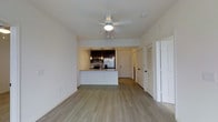 This single-floor residence features an open-concept design that seamlessly connects the living area to the kitchen, creating a spacious and functional floor plan. The living space is finished with light-toned hardwood-style flooring that runs throughout, contributing to a clean and cohesive aesthetic. A ceiling fan with an integrated light fixture is centrally mounted, providing both airflow and ambient lighting to the main living area.

The kitchen is positioned toward the rear of the open space and is defined by dark espresso cabinetry paired with granite or stone-look countertops. Stainless steel appliances, including a refrigerator and microwave, add a modern touch. Pendant lighting hangs above the kitchen peninsula, which also serves as a casual dining or prep surface and helps delineate the kitchen from the living area.

White walls throughout the unit keep the interior bright and neutral. Several doors are visible, including a louvered closet door and what appears to be an entry door, suggesting practical storage solutions are integrated into the layout. The overall ambiance is bright, understated, and move-in ready, suited for everyday living.