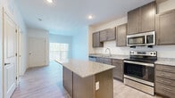 This thoughtfully designed single-floor residence offers a seamless blend of modern finishes and functional living spaces. The kitchen serves as the heart of the home, showcasing rich, dark-toned shaker-style cabinetry, granite countertops with subtle speckled detailing, and a full suite of stainless steel appliances including a refrigerator, range, built-in microwave, and dishwasher. A center island with matching granite provides additional prep space and adds to the open, airy feel of the layout. Light wood-style flooring runs throughout the main living areas, complementing the neutral color palette and recessed lighting overhead. The living area extends naturally from the kitchen, with a sliding glass door or large window bringing in generous natural light and views of the surrounding greenery. The bedrooms feature plush wall-to-wall carpeting and bright windows that frame pleasant outdoor scenery, creating a calm and restful atmosphere. The bathroom is finished with consistent design elements, including a granite-topped vanity with a undermount sink, dark wood cabinetry, a large mirror, and a shower-tub combination, all set against clean white walls and neutral tile flooring. The overall aesthetic is cohesive, contemporary, and move-in ready.