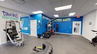 This fitness facility is a single-floor gym designed to support a wide range of workout styles and training goals. The space is organized into distinct functional zones, all connected within an open, spacious floor plan finished with durable interlocking rubber flooring throughout.

The cardio zone features a lineup of treadmills, elliptical trainers, and a stationary cycling bike, positioned alongside windows fitted with horizontal blinds that allow natural light to filter in. A wall-mounted flat-screen TV is conveniently placed for entertainment during workouts.

The strength and free weights area includes a multi-tiered dumbbell rack with a full range of weights, an adjustable bench, and a cable machine for versatile resistance training. Functional training equipment is prominently featured, including a large tire, a tire flip sled, a heavy punching bag, a multi-ball storage rack with medicine and stability balls, and plyo boxes.

Bold, vibrant blue accent walls serve as a defining design element, adorned with large motivational phrases such as "Never Quit!" and "Train Hard, Push Yourself," creating an energizing atmosphere. The dropped ceiling with recessed panel lighting ensures consistent, even illumination throughout.

Multiple doorways connect to additional areas, and the overall layout provides ample open floor space for dynamic movements, group training, or functional fitness circuits. Safety features including fire alarm systems and an AED sign are also present throughout the facility.