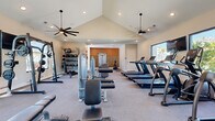 Located at 109 Pygmy Date Blvd in Venice, Florida, this single-story fitness center is thoughtfully designed to accommodate a full range of workout activities. The main exercise floor is anchored by soaring vaulted ceilings fitted with large ceiling fans and recessed lighting, creating an airy, open environment that encourages movement. Carpet flooring throughout provides comfort underfoot, while oversized windows along the perimeter flood the space with natural light and offer views of the surrounding outdoor landscape.

The main workout area is comprehensively outfitted with a row of treadmills, elliptical machines, and cable and strength training stations, along with a full rack of dumbbells displayed alongside a large wall mirror — ideal for form monitoring during free weight sessions. A dedicated medicine ball and accessory rack rounds out the strength training offerings.

A separate spin room features warm accent walls in an earthy terracotta tone, complemented by textured wood wall paneling, and houses stationary cycling bikes for focused cardio sessions. The entry area includes a posted fitness rules board and sanitation station, reinforcing a clean and organized atmosphere. Overall, the facility presents a modern, well-maintained environment designed to support diverse fitness goals in a bright and comfortable setting.