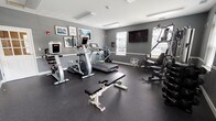 The Kensington's fitness center is a thoughtfully designed amenity space that provides residents with a well-rounded workout environment. The room features neutral gray walls accented by white trim and baseboards, creating a clean, modern aesthetic throughout. Durable rubber flooring covers the entire space, providing both safety and functionality for a variety of exercises.

The fitness center is divided into two connected areas. The primary cardio zone houses a treadmill, elliptical, and stationary bike, all from reputable equipment brands. A flat-screen television is wall-mounted nearby, and motivational framed artwork decorates the walls. A water station with a floor-standing cooler and supply cart ensures hydration is readily accessible.

The adjoining strength training area features a multi-station cable machine, a set of free-weight dumbbells with a dedicated rack, and an adjustable weight bench. A large wall mirror spans one side of this section, visually expanding the space and allowing users to monitor their form. Natural light filters in through several windows fitted with blinds, and a glass-paneled door provides direct exterior access. Overhead fluorescent lighting ensures the space remains well-lit throughout the day.