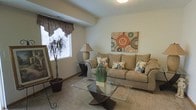 This two-story residence offers a practical and comfortable layout spread across two floors. The living room, pictured here, serves as a central gathering space characterized by soft carpeted flooring and neutral-toned walls that create a warm, inviting atmosphere. A window dressed with a decorative valance allows natural light to filter into the room, brightening the space and providing a connection to the outdoors. The room comfortably accommodates a full-sized sofa with accent pillows, a glass-top coffee table, and decorative elements including potted plants and wall art, demonstrating its generous proportions. Subtle architectural details such as a structural beam and a textured ceiling contribute to the room's character. The overall design of the living area leans toward a relaxed, residential aesthetic with light-colored walls and understated finishes that provide a neutral backdrop adaptable to various décor styles. With two floors, this residence offers distinct zones for living and other functions, making it a versatile space suited for everyday use.