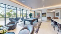 This 1,070 square foot clubroom in Miami, FL, exemplifies modern luxury and versatility. The space is characterized by its open-concept design, featuring high ceilings with recessed lighting and LED strips that create a warm, inviting ambiance. Large windows allow ample natural light and offer views of the surrounding area.

The main lounge area is furnished with a plush leather sofa and complementary seating, centered around a striking abstract artwork. A sleek kitchenette boasts marble countertops, stainless steel appliances, and ample cabinetry, perfect for entertaining. The space seamlessly transitions to a dining area with a large island and modern bar stools.

A game room adds a playful touch with a pool table, surrounded by comfortable seating and more vibrant artwork. Throughout the space, carefully curated decor elements, including potted plants and contemporary light fixtures, enhance the sophisticated atmosphere.

The flooring alternates between light-colored tiles and patterned area rugs, adding texture and defining different functional zones. The overall color palette of neutrals with pops of color in the furnishings and art pieces creates a cohesive and stylish environment.

This clubroom is designed to cater to various activities, from casual gatherings to more formal events, making it an ideal communal space for residents or guests to relax, socialize, and entertain.