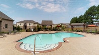 This outdoor amenity area centers around a generously sized freeform swimming pool with clear turquoise water, framed by a textured aggregate deck with classic brick border detailing. Entry steps with handrails provide easy access, and depth markers are clearly visible along the pool's edge. The surrounding deck accommodates multiple lounge chairs and seating arrangements, offering ample space for relaxation.

A covered pergola structure serves as a shaded retreat adjacent to the pool, furnished with woven lattice chairs and cast metal tables, providing a comfortable outdoor gathering spot. A colorful umbrella adds a casual accent to the poolside atmosphere. The entire area is enclosed by black decorative iron fencing, providing security while maintaining an open, airy feel.

Mature evergreen shrubs and carefully maintained landscaping beds line the perimeter, adding greenery and natural privacy. Views of the surrounding residential buildings and open grassy areas are visible beyond the fence line. The overall ambiance is clean, well-maintained, and inviting, designed to offer a relaxing outdoor experience in a community setting.