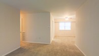 This single-floor space offers an open and functional layout designed for comfortable everyday living. The interior is finished with wall-to-wall neutral-toned carpeting throughout, creating a cohesive and understated aesthetic across the living areas. Smooth, cream-colored walls provide a clean backdrop, complemented by white baseboards that frame each room neatly.

A partial interior wall divides the space, adding subtle definition between areas while maintaining an open feel. One section of the space is anchored by a ceiling fan with a built-in light fixture, offering both air circulation and ambient lighting. A window in this area allows natural light to filter in, brightening the room and contributing to an airy atmosphere.

Additional functional details include a wall-mounted thermostat for climate control and a ventilation grille positioned low on the wall, indicating a forced-air HVAC system. The overall design is straightforward and practical, with a neutral palette that provides a versatile foundation for a variety of furnishing styles. The single-floor layout ensures easy accessibility throughout the space.