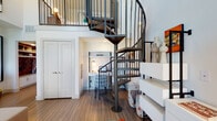 This thoughtfully designed loft-style space combines modern aesthetics with functional living. At its centerpiece stands a dramatic steel spiral staircase that connects the main living area to an elevated loft above, adding vertical dimension and architectural character to the open floor plan. The living area features warm wood-style flooring, generous natural light from slatted window treatments, and ample room for comfortable seating and lounging.

The kitchen is well-equipped with stainless steel appliances — including a refrigerator, electric range, microwave, and dishwasher — paired with crisp white shaker-style cabinetry extending to the ceiling for abundant storage, and granite-style countertops. The seamless flow between the kitchen and living area makes the space feel open and connected.

The bedroom is a calm, clean retreat with warm flooring and bright white walls that provide a neutral canvas. The bathroom features a granite-topped double vanity with white cabinetry, a tub-shower combination with tile surround, and wood-style flooring that ties the space together. A dedicated workspace nook tucked beneath the loft level adds versatility and practicality, making this space well-suited for both living and working from home.