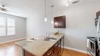 This thoughtfully designed 1-bedroom, 1-bathroom residence offers a functional and cohesive layout across a single floor. The kitchen serves as a standout feature, showcasing rich dark-wood cabinetry, granite-style countertops, and a full suite of stainless steel appliances including a refrigerator, range, and dishwasher. A peninsula counter with pendant lighting above creates a natural division between the kitchen and the open living area, enhancing both the workflow and the overall sense of space. Warm hardwood-style flooring runs consistently throughout the unit, lending a unified and inviting aesthetic. The living area benefits from a ceiling fan and an open, airy feel accentuated by neutral gray walls and clean white trim. The bedroom is a comfortable, well-proportioned room with the same hardwood-style flooring, a ceiling fan, and convenient access to a dedicated closet. The bathroom is cleanly appointed, featuring a full tub-shower combination with a curtain enclosure, a toilet, and a vanity. A washer unit is also visible near the bathroom, adding practical in-unit laundry convenience. Overall, the space maintains a clean, modern, and functional character suited to everyday living.
