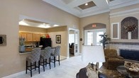 This single-story space showcases a well-appointed interior with warm beige walls and light hardwood-style flooring throughout. The main area features a coffered ceiling with recessed lighting, adding architectural depth and a sense of height to the open layout. A kitchen or kitchenette area is positioned to one side, equipped with wood-toned cabinetry, a black refrigerator, and a breakfast bar lined with patterned upholstered bar stools, making it suitable for casual gatherings or events. The open lounge area is furnished with comfortable seating and accented by a distinctive natural wood lamp base, contributing to a relaxed yet refined atmosphere. Arched doorways and French-style glass doors with white framing provide elegant transitions between spaces, allowing natural light to filter through from exterior entry points. A glimpse into an adjoining room reveals additional seating, suggesting multiple functional areas within the layout. Decorative framed artwork and lush potted plants add finishing touches that enhance the overall inviting ambiance. The space appears designed for communal or social use, combining practicality with tasteful design elements.