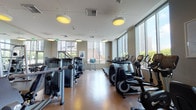 Located at 225 S Grand Ave in the heart of downtown Los Angeles, this fitness center offers a comprehensive workout environment across a single, spacious floor. The facility is defined by its floor-to-ceiling windows that line the perimeter, flooding the space with natural light and providing sweeping views of the downtown Los Angeles skyline, creating an energizing backdrop for exercise.

The cardio section is generously appointed with a full row of treadmills, elliptical trainers, stair climbers, and stationary bikes — including Life Fitness branded equipment — each unit positioned to take advantage of the city views. A dedicated strength training area houses a wide array of cable machines, multi-function cable systems, and selectorized weight machines suited for full-body workouts. A free weight zone features a complete dumbbell rack alongside a rubber-floored mat area for functional and flexibility training, complemented by stability balls.

Large mirrors line sections of the interior walls, enhancing the sense of space and supporting proper form during workouts. The flooring throughout is a durable, warm-toned hardwood-style surface, transitioning to rubber matting in the free weight area. Overhead pendant lighting and recessed fixtures provide consistent, even illumination. Framed artwork on the walls and a mounted flat-screen television add polished, modern finishing touches to this well-rounded urban fitness facility.