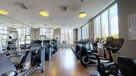 Located at 225 S Grand Ave in the heart of downtown Los Angeles, this fitness center offers a comprehensive workout environment across a single, spacious floor. The facility is defined by its floor-to-ceiling windows that line the perimeter, flooding the space with natural light and providing sweeping views of the downtown Los Angeles skyline, creating an energizing backdrop for exercise.

The cardio section is generously appointed with a full row of treadmills, elliptical trainers, stair climbers, and stationary bikes — including Life Fitness branded equipment — each unit positioned to take advantage of the city views. A dedicated strength training area houses a wide array of cable machines, multi-function cable systems, and selectorized weight machines suited for full-body workouts. A free weight zone features a complete dumbbell rack alongside a rubber-floored mat area for functional and flexibility training, complemented by stability balls.

Large mirrors line sections of the interior walls, enhancing the sense of space and supporting proper form during workouts. The flooring throughout is a durable, warm-toned hardwood-style surface, transitioning to rubber matting in the free weight area. Overhead pendant lighting and recessed fixtures provide consistent, even illumination. Framed artwork on the walls and a mounted flat-screen television add polished, modern finishing touches to this well-rounded urban fitness facility.