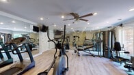Located at 2 Hollandale Lane in Clifton Park, NY, this well-appointed fitness center offers a comprehensive workout environment on a single floor. The main exercise area is generously sized, featuring light hardwood-style flooring, crown molding, and recessed lighting throughout, creating a clean and polished atmosphere. A ceiling fan with a light fixture provides added comfort during workouts. Full-length wall mirrors amplify the sense of space and allow users to monitor their form. The facility is equipped with multiple treadmills, elliptical machines, a multi-station cable and strength system, and a flat bench, offering a well-rounded selection of cardio and resistance training options. A large window on one wall invites natural light and provides a view of the surrounding greenery. Adjacent to the main workout floor is a changing and shower area, featuring multiple curtained shower stalls with clean, neutral finishes. The restroom includes a dual-sink vanity with a marble-style countertop, framed mirrors, and matching hardwood-style flooring, maintaining a consistent and refined aesthetic throughout the facility. Emergency exit signage and safety features are also in place, underscoring the facility's attention to detail and user well-being.