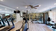 Located at 2 Hollandale Lane in Clifton Park, NY, this well-appointed fitness center offers a comprehensive workout environment on a single floor. The main exercise area is generously sized, featuring light hardwood-style flooring, crown molding, and recessed lighting throughout, creating a clean and polished atmosphere. A ceiling fan with a light fixture provides added comfort during workouts. Full-length wall mirrors amplify the sense of space and allow users to monitor their form. The facility is equipped with multiple treadmills, elliptical machines, a multi-station cable and strength system, and a flat bench, offering a well-rounded selection of cardio and resistance training options. A large window on one wall invites natural light and provides a view of the surrounding greenery. Adjacent to the main workout floor is a changing and shower area, featuring multiple curtained shower stalls with clean, neutral finishes. The restroom includes a dual-sink vanity with a marble-style countertop, framed mirrors, and matching hardwood-style flooring, maintaining a consistent and refined aesthetic throughout the facility. Emergency exit signage and safety features are also in place, underscoring the facility's attention to detail and user well-being.