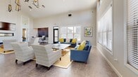 Located at 4001 De Morada Dr in Odessa, TX, this single-floor open-concept space offers a well-appointed layout combining a living room and kitchen in one cohesive, flowing environment. The soaring ceilings—dramatically accentuated by clerestory windows positioned high on the walls—invite abundant natural light throughout the day, creating a bright and airy atmosphere.

The living area is expansive, featuring large windows dressed with horizontal blinds that offer views of the surrounding neighborhood. The space is anchored by tile flooring, a wall-mounted TV above a sleek linear fireplace, and a built-in workspace nook with a wood-paneled accent wall. A separate seating zone with blue sofas and yellow accent pillows adds a vibrant, contemporary feel. Glass-panel doors connect interior zones and extend the sense of openness.

The kitchen is a standout feature, distinguished by dramatic floor-to-ceiling stacked stone accent walls that lend a rustic yet refined character. White cabinetry pairs with granite countertops, stainless steel appliances, and floating reclaimed wood shelves. A central island with bar seating and pendant lighting above completes the kitchen, making it both functional and visually striking throughout this well-designed space.