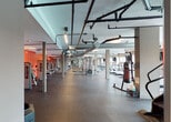 Located at 6350 Griffin Rd in Davie, Florida, this single-floor fitness center offers a comprehensive and well-equipped workout environment designed to accommodate a wide range of training styles. The space is organized into distinct functional zones, each thoughtfully arranged to maximize usability and flow.

The cardio area features a generous row of treadmills, ellipticals, stationary bikes, and stair climbers, positioned alongside large floor-to-ceiling windows that invite natural light and offer views of the surrounding outdoor landscape. Mounted televisions are positioned throughout for convenience during workouts.

The strength training section is expansive, housing an extensive collection of dumbbells on multiple racks, weight benches, barbells, and a full lineup of cable and resistance machines. Bold red accent walls energize this zone, creating a dynamic and motivating atmosphere. Full-length mirrors line the walls, enhancing both the sense of space and functional utility for form-checking.

Overhead, exposed industrial-style ductwork, pipes, and ventilation systems run across the ceiling, lending the space a modern, urban aesthetic. The flooring transitions between smooth tile in open corridors and durable rubber matting in the free weights and equipment areas, providing appropriate surfaces for varied training activities. Structural columns are integrated throughout, defining zones while maintaining an open, airy feel across the entire facility.
