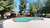 This single-story community amenity space offers a welcoming and functional outdoor-oriented environment set among mature, leafy trees. At the heart of the property is a generously sized octagonal outdoor swimming pool with a clean, teal-blue finish and rust-accented border, surrounded by a spacious concrete pool deck ideal for lounging. The pool area is enclosed by a wrought iron fence and offers ample room for sunbathing and relaxation.

The clubhouse building features a covered entrance porch supported by white columns, outfitted with recessed lighting and large windows that look out directly toward the pool — creating a seamless connection between interior and exterior spaces. French doors with glass panes provide an inviting entryway into the building. The covered patio also includes outdoor table-and-chair seating, offering a shaded spot to relax just steps from the water.

Adjacent to the pool area, a separate outdoor patio features additional seating beneath a vibrant red umbrella, with views extending to well-maintained green lawns, tennis courts, and a small gazebo structure in the distance. Floodlights positioned throughout the grounds suggest extended usability into the evening hours. The entire space is framed by lush, mature trees and manicured landscaping, contributing to a peaceful, park-like atmosphere throughout the community.