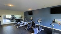 This fitness center is a dedicated, single-floor exercise facility designed to meet a variety of workout needs. The space features a dark rubberized flooring, ideal for high-impact activities and equipment stability. A striking deep blue-grey accent wall serves as the room's focal point, anchoring a wall-mounted flat-screen TV — perfect for entertainment during cardio sessions. The cardio equipment lineup includes multiple stationary bikes, both upright and recumbent styles, as well as elliptical trainers, providing options for low-impact and full-body cardiovascular workouts. A full mirrored wall runs along one side of the space, visually expanding the room and allowing users to monitor their form during exercise. A well-stocked dumbbell rack is positioned in front of the mirror, offering a range of free weights for strength training. The ceiling is smooth and neutral, complementing the clean, modern aesthetic of the room. The overall ambiance is functional and focused, with a layout that encourages efficient use of space while maintaining an open, uncluttered feel throughout the facility.