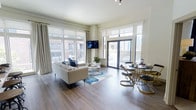 This thoughtfully designed single-floor space offers a seamless blend of modern style and functional living. The open-concept layout connects the living and dining areas in a fluid, airy arrangement that maximizes the sense of space throughout.

The living area is anchored by rich hardwood flooring that extends across the entire floor, complemented by crisp white walls that enhance the brightness of the room. An abundance of natural light pours in through large, floor-to-ceiling windows fitted with roller shades, offering both sun control and privacy while preserving the connection to the outdoors. A wall-mounted flat-screen television serves as a focal point above the seating area.

The dining area features a sleek, round glass-top table paired with modern gold-accented chairs, positioned conveniently near the expansive glazed doors that open directly onto an outdoor balcony. The balcony provides open urban views, bringing a dynamic cityscape backdrop to the interior.

Tall ceilings amplify the sense of volume, while the neutral color palette throughout creates a clean, contemporary ambiance. Track lighting adds a refined touch to the ceiling, allowing for flexible illumination. The overall atmosphere is sophisticated yet welcoming, ideal for both everyday living and entertaining.