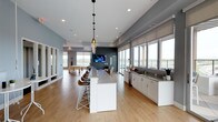 This single-floor communal space is designed with a sleek, modern aesthetic that blends functionality with an inviting atmosphere. The open-concept layout creates a seamless flow between distinct zones, making it ideal for both casual gatherings and social events. Light hardwood flooring runs throughout, complementing the cool gray and white color palette that defines the interior.

At the heart of the space is a large central island with a stone countertop and leather bar stools beneath elegant pendant lighting, providing an ideal spot for dining or conversation. Adjacent to the island, a dedicated kitchenette features white cabinetry, dark countertops, a sink, and a dishwasher — offering practical amenities for hosting.

The lounge area is anchored by a wall-mounted flat-screen TV set against a bold dark accent wall, while a pool table nearby adds a recreational dimension to the space. Natural light floods the interior through a generous row of large windows fitted with roller shades, offering panoramic outdoor views and enhancing the bright, airy ambiance. Recessed ceiling lighting provides consistent, even illumination throughout. Together, these elements create a well-appointed, versatile space suited for a variety of social and recreational activities.