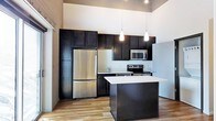 This single-floor space showcases a contemporary kitchen designed with both style and functionality in mind. The kitchen is anchored by rich, dark-finished cabinetry that extends from floor to ceiling, providing generous storage while creating a bold visual contrast against the light-toned walls and ceilings. The cabinetry layout is well-organized, featuring both upper and lower cabinets along with tall pantry-style units that maximize vertical space.

Stainless steel appliances, including a full-size refrigerator, electric range, built-in microwave, and sink, are seamlessly integrated into the design, contributing to a polished, modern aesthetic. Light-colored countertops provide a striking contrast to the dark cabinetry, offering ample workspace for meal preparation.

A central kitchen island echoes the dark cabinetry finish and serves as a natural gathering point, ideal for casual dining or additional prep space. Overhead, two elongated teardrop pendant lights hang with purpose, directing warm light over the island. The flooring features light wood-tone planks that add warmth to the space. Notably tall ceilings amplify the sense of openness throughout the kitchen, enhancing the overall airy ambiance.