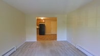 This single-floor residential space offers a straightforward and functional layout designed for comfortable everyday living. The main living area features wall-to-wall carpeting in a neutral tone, providing a warm and quiet underfoot experience. Light-colored walls throughout create a clean, bright atmosphere, enhanced by the open sightlines between rooms. A wide open passage connects the living area directly to the kitchen, promoting an easy flow between spaces. The kitchen is equipped with warm-toned wooden cabinetry providing ample storage, a black refrigerator, and hard-surface flooring that contrasts with the carpeted living space. A flush-mount ceiling light fixture illuminates the kitchen area. A door off the kitchen offers additional access, adding to the unit's practical layout. The space maintains a simple, unadorned aesthetic with white trim and baseboards framing each room cleanly. Overall, the layout is compact yet functional, well-suited for those seeking a low-maintenance living environment with clearly defined areas for daily activities.