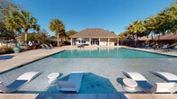 This outdoor community pool facility in Baton Rouge, Louisiana offers a well-appointed resort-style experience designed for relaxation and social gathering. The centerpiece is a generously sized swimming pool with crystal-clear blue water, complete with in-water lounge chairs and pedestals that allow residents to cool off while enjoying the Louisiana sunshine. The pool deck is expansive, lined with blue lounge chairs and shaded umbrella stations, providing ample space for sunbathing and leisure.

A covered pavilion anchors one side of the pool area, constructed with sturdy wooden beams and an open-air design. This shaded gathering space is outfitted with multiple round tables and chairs, and features a mounted flat-screen television overhead — making it an ideal venue for outdoor entertainment and socializing. Adjacent to the pavilion, a dedicated grilling station with a gas grill and picnic table offers a convenient outdoor cooking area.

The entire pool complex is enclosed by a decorative wrought iron fence, ensuring both security and an aesthetically pleasing boundary. Tall palm trees and mature landscaping frame the space, creating a tropical ambiance. A pool house with a hip roof serves as a backdrop, completing this inviting and functional outdoor amenity.