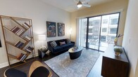 This single-floor residence showcases a thoughtfully designed living space that blends modern aesthetics with functional comfort. The living room serves as the centerpiece of the home, featuring dark hardwood-style flooring that grounds the space with warmth and sophistication. Elevated ceilings fitted with a ceiling fan contribute to an open, airy atmosphere throughout the room.

Large floor-to-ceiling sliding glass doors flood the interior with natural light while providing direct access to a private balcony, which overlooks a shared outdoor amenity area. The expansive glazing creates a seamless visual connection between the interior and exterior, enhancing the overall sense of space.

The living area is well-appointed, with ample room for a full sofa arrangement centered around a statement oval coffee table atop a geometric-patterned area rug. Bold, colorful artwork adorns the neutral-toned walls, adding character and visual interest. A contemporary floor lamp and coordinating side table complete the curated look. The overall ambiance is polished and modern, balancing vibrant decorative accents against a clean, neutral architectural backdrop.