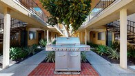 Located on Victory Boulevard in Reseda, California, this multi-unit residential community offers a well-rounded set of shared amenities designed to support an active and social lifestyle. The building's warm stucco exterior, finished in a classic tan and yellow palette, frames a central courtyard that serves as the heart of the community. At the center of the courtyard stands a mature shade tree, beneath which a large stainless steel gas grill is mounted on a red brick platform — an inviting setup for outdoor gatherings and al fresco dining. Manicured drought-tolerant landscaping with ornamental grasses and succulents lines the walkways, adding greenery and texture to the outdoor spaces. A pool area is accessible from the courtyard, bordered by iron fencing and open stairways connecting upper balcony corridors. Dedicated bicycle parking is conveniently situated near building access points. The on-site fitness center is well-equipped, featuring treadmills, ellipticals, free weights, medicine balls, and resistance equipment, with bold accent walls and wall-to-wall mirrors enhancing the workout environment. Natural light filters in through the fitness room's windows, creating a functional and energizing space. The property provides underground parking access, rounding out the practical amenities available to residents.