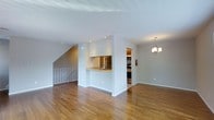 This two-floor residence offers a well-designed layout that balances open living with functional flow. The main level features warm hardwood flooring throughout, lending a sense of continuity and warmth to the space. A central architectural partition defines the kitchen area, which includes a pass-through counter with a sink, providing a seamless connection between the kitchen and the surrounding living area. The kitchen is equipped with stainless steel appliances, including a refrigerator and range, complemented by dark cabinetry for added contrast. Recessed lighting in the kitchen ensures a well-lit workspace, while an elegant chandelier in the adjacent dining or living area adds a refined decorative touch. The open-concept design of the main floor creates a spacious, airy feel, ideal for both everyday living and entertaining. A staircase with a metal railing, visible toward the rear of the main floor, provides access to the second level, where additional living or sleeping quarters are located. The white walls and clean architectural lines throughout contribute to a bright, modern aesthetic, making this a practical and visually cohesive multi-level residence.
