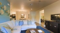 This single-story space showcases an open-concept floor plan that seamlessly connects the main living areas. The living room serves as the central gathering space, featuring recessed lighting, a ceiling fan, and warm-toned walls that create an inviting atmosphere. Wood-style flooring flows throughout, grounding the space with a clean, contemporary feel.

A spacious sectional sofa anchored by a deep blue area rug defines the living area, complemented by a round wood coffee table that adds warmth. Blue and white accent pillows introduce a fresh, casual aesthetic. Recessed lighting and a decorative pendant light overhead provide layered illumination.

The adjoining dining area is visible in the background, highlighted by a hanging light fixture and wall art, creating a defined yet connected space. The kitchen sits nearby, fitted with dark cabinetry and modern appliances, offering functional contrast to the lighter living areas.

A hallway visible to the right leads to additional rooms, hinting at further living space beyond. Natural light filters in from adjoining rooms, contributing to the bright and airy overall ambiance of this well-connected, single-level home.