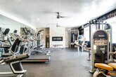 This fitness center is a thoughtfully designed exercise facility offering a comprehensive range of workout equipment and amenities across a single floor. The space is divided into distinct functional zones, each tailored to a specific type of training.

The cardio area features multiple treadmills, elliptical machines, and a stationary bike, all arranged on durable rubber flooring. A large wall mirror amplifies the sense of space in this zone, while a ceiling fan overhead provides air circulation during intense workouts. Recessed lighting and a textured ceiling complete the practical aesthetic.

The strength training area is anchored by a striking stacked-stone accent wall housing a sleek linear fireplace, creating an unexpectedly warm and inviting atmosphere. A cable machine, multi-function weight station, and adjustable bench provide versatile resistance training options. An additional cable and pulley system is positioned nearby for further variety.

Bold motivational typography covers the dark-painted accent walls throughout, featuring words such as "strength," "commitment," "dedication," and "challenge," reinforcing an energizing atmosphere. Natural light enters through glass-panel doors and windows, connecting the space to the outdoors.

A transition area with light wood-look flooring leads to clearly marked, separate men's and women's restroom facilities, adding convenience to the overall design.