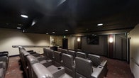 This dedicated home theater is designed to deliver a true cinema experience within a single-floor layout. The room features tiered, stadium-style seating arranged across multiple rows, ensuring unobstructed sightlines from every position. The plush, dark leather recliners are equipped with built-in cup holders and wide armrests, prioritizing comfort during extended viewing sessions.

The matte black ceiling is a defining architectural element, effectively minimizing light reflection and enhancing the immersive atmosphere of the space. Recessed ceiling lighting provides soft, controlled ambient illumination, complemented by subtle wall sconces that add warmth without compromising the viewing environment. The walls are finished in neutral, dark tones that further absorb light and reduce glare.

A large flat-screen display is mounted at the front of the room, serving as the focal point of the theater. A concession-style counter area is visible toward the rear, adding a functional and entertaining amenity to the space. The overall design balances practicality with a refined, dedicated entertainment aesthetic.