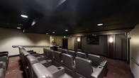 This dedicated home theater is designed to deliver a true cinema experience within a single-floor layout. The room features tiered, stadium-style seating arranged across multiple rows, ensuring unobstructed sightlines from every position. The plush, dark leather recliners are equipped with built-in cup holders and wide armrests, prioritizing comfort during extended viewing sessions.

The matte black ceiling is a defining architectural element, effectively minimizing light reflection and enhancing the immersive atmosphere of the space. Recessed ceiling lighting provides soft, controlled ambient illumination, complemented by subtle wall sconces that add warmth without compromising the viewing environment. The walls are finished in neutral, dark tones that further absorb light and reduce glare.

A large flat-screen display is mounted at the front of the room, serving as the focal point of the theater. A concession-style counter area is visible toward the rear, adding a functional and entertaining amenity to the space. The overall design balances practicality with a refined, dedicated entertainment aesthetic.