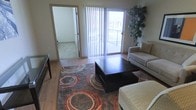 This single-floor space offers a well-organized living area centered around wood-style flooring and an eye-catching area rug with circular patterns in warm earth tones. The living room is furnished with a neutral-toned sofa and a substantial dark wood coffee table, creating a comfortable and functional gathering area. A large sliding glass door with vertical blinds opens directly onto a private balcony, allowing natural light to fill the room while providing outdoor views of the surrounding building exterior. A tall potted plant near the balcony entrance adds a natural, decorative element to the space. An open doorway connects the living area to an adjoining carpeted room, offering a clear visual transition between the spaces and highlighting the unit's straightforward, accessible layout. The overall ambiance is bright and neutral, with light-toned walls that complement the warm flooring and textured furnishings. The combination of natural light from the balcony access, the contrast between hardwood-style and carpeted flooring zones, and the open sightlines between rooms gives the space a practical and welcoming character suitable for everyday living.