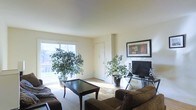 This single-floor space offers a welcoming and comfortable living environment defined by its clean lines and naturally lit interior. The living area is the centerpiece of the home, anchored by a large sliding window that floods the room with abundant natural light and offers a view of the surrounding outdoor greenery. The warm hardwood flooring extends throughout, complementing the neutral, cream-toned walls that create a bright and airy atmosphere. The space is thoughtfully arranged, accommodating a plush seating area with a dark upholstered sofa and a sleek dark wood coffee table, ideal for relaxation and entertaining. Lush indoor plants placed near the window add a vibrant, organic touch to the room's décor. A dedicated media area with a flat-screen television and slim media stand sits against one wall, framed by a tasteful piece of wall art. The high, smooth ceilings contribute to the sense of openness and spaciousness throughout. A white interior door provides access to additional areas of the home. Overall, the space presents a harmonious blend of function and comfort, with a light-filled, inviting character suitable for everyday living.