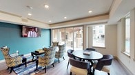 This single-floor communal lounge is a thoughtfully designed gathering space that blends functional amenities with contemporary style. The open layout is anchored by a fully equipped kitchen area featuring dark wood cabinetry, a herringbone-tiled backsplash, stainless steel appliances including a refrigerator, dishwasher, and microwave, and a large white-topped island with bar seating. Pendant lights with brass hardware hang above the island, adding warmth to the space.

The lounge area is divided into distinct zones. A TV lounge features a wall-mounted flat-screen television set against a bold teal accent wall, complemented by patterned armchairs in blue, gold, and white geometric prints arranged around a dark wood coffee table. An adjacent dining area offers a round table with upholstered chairs positioned beside large sliding glass doors and expansive windows that flood the space with natural light and provide direct views of an outdoor pool and covered patio.

Wood-look flooring runs throughout, and recessed lighting provides consistent illumination. Crown molding details add a refined finishing touch. The overall ambiance is vibrant yet inviting, combining bold color choices with practical, well-appointed furnishings suited for social gatherings.