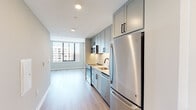 This thoughtfully designed urban residence offers a clean, contemporary aesthetic with consistent finishes throughout its single floor layout. The galley-style kitchen is a standout feature, equipped with stainless steel GE appliances including a French door refrigerator, electric range, over-the-range microwave, and dishwasher. Light gray shaker-style cabinetry with matte black hardware lines the walls from floor to ceiling, complemented by crisp white quartz countertops and a classic subway tile backsplash. A deep undermount sink with a sleek matte black faucet completes the culinary space.

Light hardwood-style flooring flows seamlessly throughout the living areas and bedroom, creating a warm and cohesive feel. The living and sleeping areas benefit greatly from expansive floor-to-ceiling windows framed in bold black, allowing abundant natural light to pour in while offering sweeping views of the surrounding urban skyline. The neutral, light gray wall tones enhance the bright, airy ambiance of each room.

The bathroom is finished with large-format white wall tiles surrounding a full soaking tub and shower combination, complemented by neutral floor tile, a clean white vanity, and recessed lighting. The overall design of the space balances modern functionality with a refined, minimalist sensibility ideal for urban living.