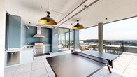This generously scaled rooftop amenity level seamlessly blends indoor and outdoor living across a substantial footprint. The interior lounge area features large-format tile flooring, exposed structural columns, and bold blue accent walls, all illuminated by striking pendant lights with gold interiors that lend a warm, contemporary glow. Floor-to-ceiling glass walls and sliding glass doors dissolve the boundary between inside and outside, flooding the space with abundant natural light and offering unobstructed panoramic views of the surrounding cityscape.

The outdoor kitchen is outfitted with a professional-grade stainless steel grill, a stainless range hood, granite countertops, and a prep sink, all accented by a stylish blue subway tile backsplash — ideal for entertaining. Adjacent to the kitchen, a full-size ping-pong table anchors the recreation area, complemented by clusters of modern wicker and molded seating arranged around circular tables throughout the lounge.

The open-air terraces extend the space further, featuring glass-panel railings that preserve uninterrupted skyline views. One terrace offers comfortable teak lounge furniture with soft cushions, while a separate sun deck is lined with vibrant chaise lounges beneath a slatted wood pergola, perfect for relaxing and taking in far-reaching urban vistas.