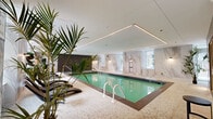 Located at 1770 Rue Joseph Manseau in Montréal, this upscale indoor aquatic and wellness facility spans a generous single floor, offering a refined retreat defined by high-end materials and thoughtful design. The centerpiece is a large indoor lap pool with crystal-clear turquoise water, bordered by sleek dark granite edging and a mosaic penny-tile surround that adds texture and warmth underfoot. Floor-to-ceiling marble-clad walls in a soft white-and-grey veining pattern lend a spa-like grandeur to the space, complemented by natural light filtering in through street-facing windows. Recessed linear LED lighting traces the ceiling perimeter, casting a warm, ambient glow that enhances the serene atmosphere. Along one side of the pool, a row of modern dark-wood lounge chairs invites relaxation, while tropical potted palms introduce a touch of organic vitality. A dedicated hot tub with hydrotherapy jets is seamlessly integrated into the layout, offering contrast to the main pool. A separate sauna room enclosed in glass and warm wood framing provides an additional wellness retreat. The combination of marble, dark slate tile, and polished mosaic flooring throughout creates a cohesive, luxurious aesthetic that positions this facility as a premium amenity space.
