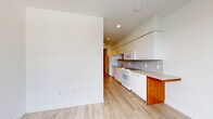 This efficiently designed single-floor unit offers a thoughtful layout that maximizes every square foot of its compact footprint. The entry is immediately distinguished by a bold red door, providing a striking accent against the crisp white walls that carry throughout the entire space. Light wood-toned vinyl plank flooring unifies all areas, lending warmth and visual continuity.

The kitchen runs along one wall in a galley-style configuration, featuring white cabinetry extending to the ceiling for ample storage, a mosaic tile backsplash, a built-in range and microwave, a refrigerator, and a countertop peninsula with a warm wood base — ideal as a breakfast bar or prep extension. Recessed lighting overhead keeps the kitchen bright and functional.

A central hallway connects the main living area to supporting spaces, with a stacked in-unit washer and dryer conveniently tucked into the corridor, alongside a dedicated closet with wire shelving for storage.

The living area is open and light-filled, benefiting from a window that draws in natural light and offers an outward view. The clean, minimalist walls provide a versatile blank canvas.

The bathroom is simply appointed with wood-look flooring, white walls, and essential fixtures, maintaining the clean aesthetic found throughout the unit.