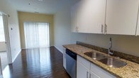 This single-floor space showcases a well-designed open layout that connects the kitchen directly to the living area, creating a seamless flow throughout. The kitchen is outfitted with rich granite countertops featuring warm brown and gold tones, contrasted by crisp white cabinetry equipped with sleek bar-pull hardware. Upper and lower cabinets provide ample storage, while a stainless steel sink and a built-in dishwasher add functional convenience. The dark hardwood flooring runs consistently throughout the space, grounding the design with a warm, polished aesthetic. Soft yellow accent walls in the living area add a welcoming touch of color, complementing the neutral gray tones of the kitchen walls. Natural light plays a prominent role in the space, streaming in generously through a large sliding glass door or wide window fitted with vertical blinds at the far end of the living area. This bright focal point creates an airy, open ambiance and hints at outdoor access. The overall design balances practicality with style, making the space both functional and visually appealing.