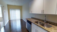 This single-floor space showcases a well-designed open layout that connects the kitchen directly to the living area, creating a seamless flow throughout. The kitchen is outfitted with rich granite countertops featuring warm brown and gold tones, contrasted by crisp white cabinetry equipped with sleek bar-pull hardware. Upper and lower cabinets provide ample storage, while a stainless steel sink and a built-in dishwasher add functional convenience. The dark hardwood flooring runs consistently throughout the space, grounding the design with a warm, polished aesthetic. Soft yellow accent walls in the living area add a welcoming touch of color, complementing the neutral gray tones of the kitchen walls. Natural light plays a prominent role in the space, streaming in generously through a large sliding glass door or wide window fitted with vertical blinds at the far end of the living area. This bright focal point creates an airy, open ambiance and hints at outdoor access. The overall design balances practicality with style, making the space both functional and visually appealing.
