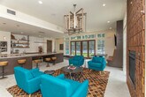 Located at 3421 N Druid Hills Rd in Decatur, Georgia, this single-floor clubhouse space blends contemporary design with warm, inviting elements to create a versatile gathering destination. The open-concept layout is anchored by a striking lounge area furnished with vibrant teal accent chairs arranged atop a richly patterned geometric rug, centered around a bold floor-to-ceiling textured brick fireplace that serves as the room's focal point. A sculptural geometric chandelier overhead adds an artistic touch to the space. Large wood-framed French doors and an abundance of transom windows flood the interior with natural light while offering expansive views of the outdoor pool area and surrounding lush greenery, seamlessly connecting the interior to the exterior. The bar and kitchen area features a generously sized granite-topped island with dark wood cabinetry, dual tap beer dispensers, open industrial-style shelving against a white subway tile backsplash, and built-in appliances — ideal for entertaining. A built-in banquette with leather upholstery and an adjacent media wall with a mounted flat-screen TV provide additional lounge and viewing options. Light-toned tile flooring flows throughout, tying the space together with a clean, cohesive aesthetic that balances modern sophistication with casual comfort.