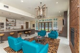 Located at 3421 N Druid Hills Rd in Decatur, Georgia, this single-floor clubhouse space blends contemporary design with warm, inviting elements to create a versatile gathering destination. The open-concept layout is anchored by a striking lounge area furnished with vibrant teal accent chairs arranged atop a richly patterned geometric rug, centered around a bold floor-to-ceiling textured brick fireplace that serves as the room's focal point. A sculptural geometric chandelier overhead adds an artistic touch to the space. Large wood-framed French doors and an abundance of transom windows flood the interior with natural light while offering expansive views of the outdoor pool area and surrounding lush greenery, seamlessly connecting the interior to the exterior. The bar and kitchen area features a generously sized granite-topped island with dark wood cabinetry, dual tap beer dispensers, open industrial-style shelving against a white subway tile backsplash, and built-in appliances — ideal for entertaining. A built-in banquette with leather upholstery and an adjacent media wall with a mounted flat-screen TV provide additional lounge and viewing options. Light-toned tile flooring flows throughout, tying the space together with a clean, cohesive aesthetic that balances modern sophistication with casual comfort.