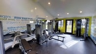 This single-floor fitness center is designed to inspire and energize its users from the moment they walk in. The space features vaulted ceilings with recessed lighting that create an open, airy atmosphere, while large glass doors and expansive windows flood the interior with natural light and offer views of the surrounding greenery outdoors.

The room is anchored by a multi-station cable and weight machine system at its center, providing a comprehensive range of strength-training options. Additional equipment, including benches and free weight accessories, is thoughtfully arranged to maximize usability and traffic flow throughout the space.

The interior design is bold and motivating, with vibrant lime-green accent walls contrasting sharply against the dark rubber flooring — a practical, durable surface ideal for a high-performance workout environment. A striking typographic mural covers one wall, featuring motivational words and phrases that reinforce a culture of commitment and achievement.

A wall-mounted split air conditioning unit ensures comfortable temperatures year-round, making the space suitable for intense workouts in any season. The combination of professional equipment, energetic design elements, and thoughtful layout makes this fitness center a dynamic and functional destination for users of all fitness levels.