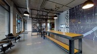 This single-floor commercial space showcases a striking industrial aesthetic defined by exposed concrete ceilings, visible ductwork, and an open network of overhead pipes and conduit. The polished concrete floors reflect light throughout, contributing to a clean yet raw industrial character. Dramatic pendant lights with dark metal shades hang from the ceiling, casting warm, focused illumination over the main communal area.

A centerpiece of the space is a long, sturdy communal table crafted from warm-toned natural wood with a dark steel frame, accompanied by matching steel-and-wood benches — ideal for collaborative gathering or casual work. Nearby, a bold graphic wall mural featuring a city map adds a strong sense of local identity and visual interest to the space.

Ornate laser-cut metal screens serve as decorative partitions, dividing areas while allowing sightlines to flow through their intricate patterned cutouts. Beyond the screens, a glimpse into an adjacent area reveals additional seating and greenery, suggesting flexible, multi-use zones within the layout. A digital display and a water station are also present, pointing to a functional, amenity-equipped environment. The overall ambiance is contemporary and purposeful, blending raw industrial materials with thoughtful design details.