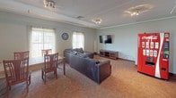 This community room is a well-appointed, single-level shared gathering space designed to accommodate residents for relaxation, socializing, and casual dining. The main lounge area features a large, dark leather sectional sofa arranged around a wall-mounted flat-screen television on a media console, creating a comfortable entertainment hub. Sheer curtains frame multiple windows that allow generous natural light to fill the room, complemented by track lighting overhead. A textured ceiling and soft neutral wall tones contribute to a relaxed, welcoming ambiance throughout.

Adjacent to the lounge, a dining area offers a table with wooden chairs, ideal for group meals or casual gatherings. The kitchenette is equipped with warm oak cabinetry, a double-basin sink with dark countertops, upper storage cabinets, and a microwave — providing convenient amenities for hosting events or everyday use. A Coca-Cola vending machine adds a practical touch near the entry.

One of the standout features of this community room is a large sliding glass door that opens directly to an indoor pool and patio area, visible through the glass and outfitted with lounge chairs and a patio umbrella. This seamless indoor-outdoor connection enhances the room's functionality and appeal as a central hub for community activity.