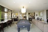 This well-appointed single-floor community space is designed with a contemporary aesthetic that balances comfort and functionality. The main lounge area is a central gathering point, furnished with generously sized sectional sofas, a wall-mounted flat-screen television atop a dark wood credenza, and soft neutral carpeting that runs throughout. Large floor-to-ceiling glass windows and doors flood the interior with abundant natural light while offering clear views of the surrounding landscaped grounds and mature trees beyond. A billiards table positioned near the windows adds a recreational element to the space.

Adjacent to the lounge, a kitchenette area features sleek white countertops with bar seating, dark cabinetry, stainless steel appliances, and a bold green accent wall adorned with a trio of framed botanical artwork. Striped sheer curtains frame the windows, softening the modern palette with a touch of warmth.

Near the entrance, floor-to-ceiling glass walls and double glass doors create a bright, welcoming entryway with small café-style seating. A glimpse of a private office or business center is also visible, enclosed by glass partitions. The overall ambiance is polished, inviting, and purposefully designed for both relaxation and community engagement.